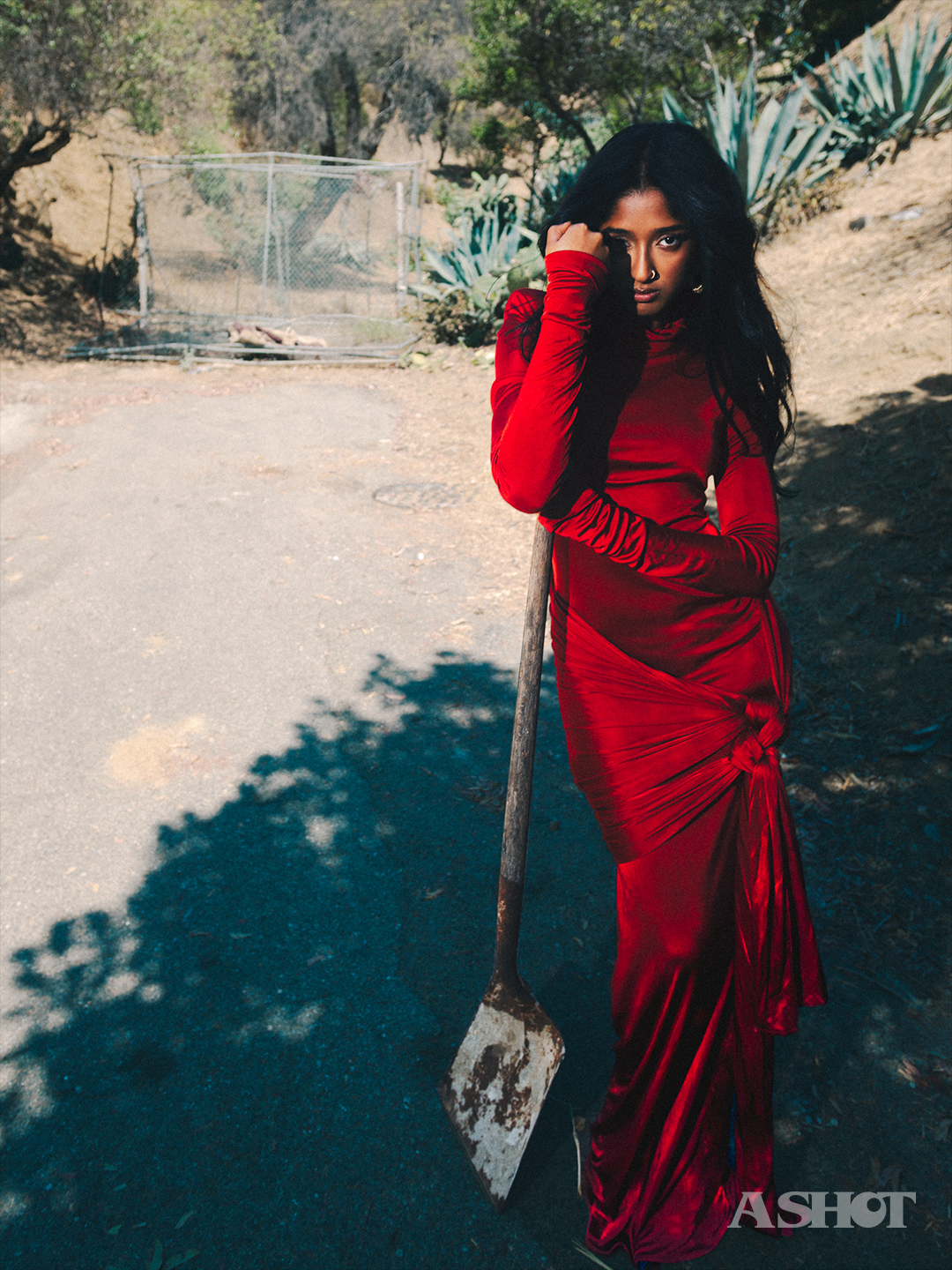 Maitreyi Ramakrishnan in a red dress leaning against a shovel against a hillside backdrop